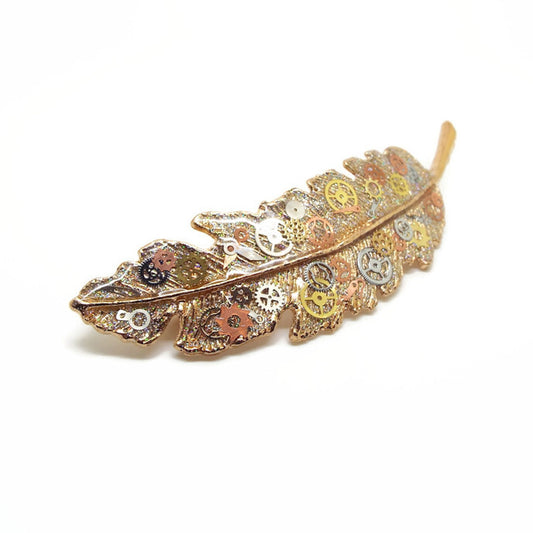 Gold coloured metal feather hair clip which is decorated with small watch parts in silver, gold and copper on a base of golden glitter with layers of clear resin