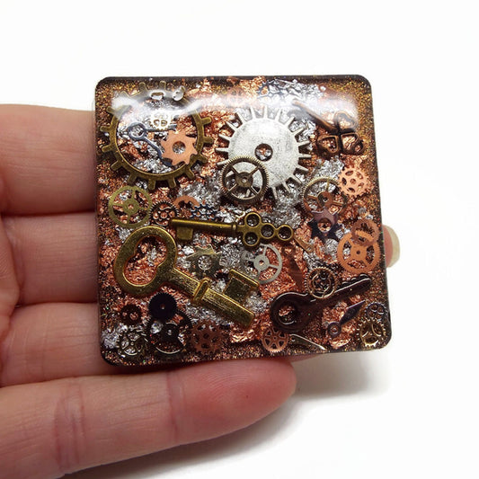 hand holding a square fridge magnet which is handmade in layers of resin with added cogs, keys and small watch parts on a background of copper and silver metallic foils and a base coat of gold glitter