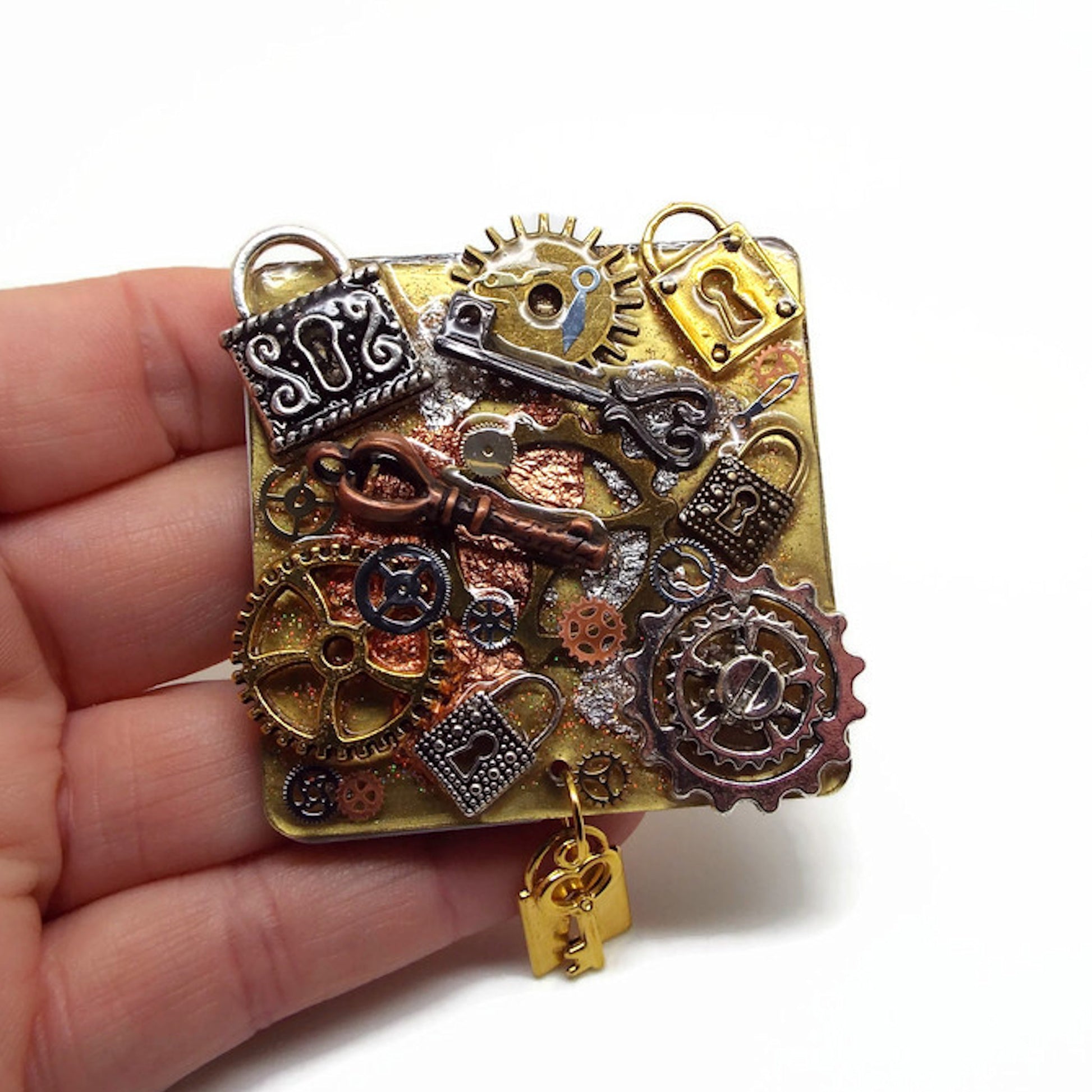 A hand holding a square fridge magnet with a gold background.  Resin has been used to attach cogs, keys, padlocks and small watch parts atop of silver and copper metallic leaf. At the bottom centre is a jump ring holding a small gold padlock and gold key